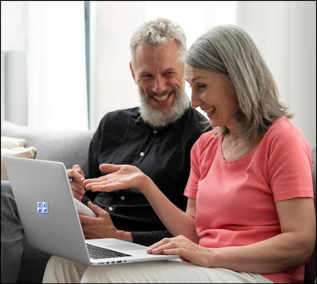 A mature woman and man sit next to each other on a couch, both smiling happily, reviewing a laptop showing their very positive retirement planning results.